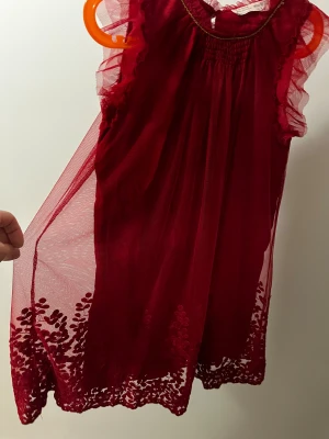 Röd tyllklänning från Zara Girls - Supersöt röd klänning från Zara Girls med volangärmar och guldfärgad detalj vid halsen. Klänningen har ett lager av tyll och broderade blommor längst ner, samt smock vid halsringningen. Perfekt för dig som gillar romantisk och lekfull stil.