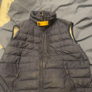 Parajumper väst - Snygg quiltad dunväst från Parajumpers i mörkblått med beige insida. Västen har hög krage med gul detalj, flera dragkedjefickor och en cool orange krok vid nacken. Perfekt för lager-på-lager och chill stil. Har ett litet håll längst nere vid fickan, går att fixa enkelt men själv ej gjort de pga ork och ingen användning av den. 
