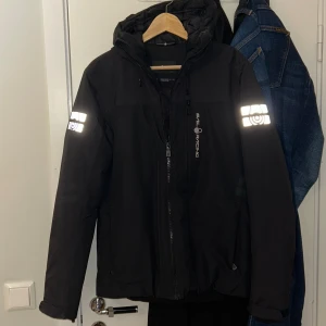Svart vinterjacka från Sail Racing - Svart winterjacka från Sail Racing med reflekterande detaljer på ärmarna och logga på bröstet. Jackan har huva med snörning, dragkedja framtill och justerbara ärmslut. Perfekt för kalla dagar och snygg streetstil. Köpte den för ungefär 3000 kr. Helt ny bestälde den men fel storlek, kunde it skicka tillbaka den så försöker sälja den här nu .