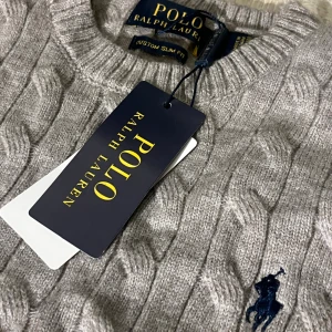 Grå kabelstickad tröja Raffe tröja  - Snygg grå kabelstickad tröja från Raffe med klassisk rund hals och marinblå broderad logga på bröstet. Tröjan har långa ärmar och ribbade muddar vid ärmslut och nederkant. Perfekt för dig som gillar stilrena och tidlösa plagg.