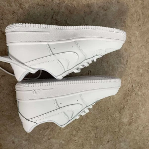 Nike Air Force 1 vita sneakers - Fräscha Nike Air Force 1 sneakers i helvitt skinn med klassisk rund tå och platt sula. Snygga detaljer som perforeringar på tån och ikoniska swoosh-loggan på sidan. Skorna har snörning och är perfekta för dig som gillar stilrena och tidlösa sneakers.