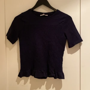 Mörkblå T-shirt med volang från Gina Tricot - Supersöt mörkblå topp från Gina Tricot med korta ärmar och volangkant nertill. Toppen har smockad struktur på framsidan som ger en cool och trendig look. Perfekt att styla med jeans eller kjol för en chill vibe.