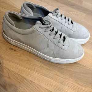 Grå sneakers från Axel Arigato i mocka - Snygga grå sneakers från Axel Arigato med stilren design och låg profil. Skorna är tillverkade i mjuk mocka med matchande grå snörning och vit platt sula. Perfekta för dig som gillar minimalistisk stil och vill ha ett par schyssta sneakers i garderoben. Använda ett fåtal gånger