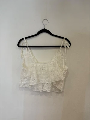 Vit croppad linnetopp med volang - Supersöt vit croppad linnetopp med broderade hålmönster och volangdetaljer. Toppen har smala axelband med knyt och en luftig, omlottliknande passform. Perfekt för varma dagar och ger en romantisk vibe till din outfit.