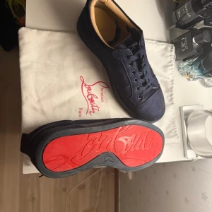 Mörkblå sneakers från Christian Louboutin - Snygga mörkblå sneakers från Christian Louboutin med klassisk röd sula och rund tå. Skorna är gjorda i mocka med svarta snören och har en stilren, minimalistisk design. Insidan är beige och sulan har Louboutins signatur. Perfekt för dig som gillar exklusiva detaljer.