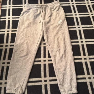 Grå mjukisbyxor från New Balance M - Grå mjukisbyxor från New Balance med elastisk midja och snörning. Byxorna har en avslappnad passform, muddar vid bensluten och logga på ena benet. Perfekta för chill eller träning, tillverkade i mjukt bomullsmaterial.
