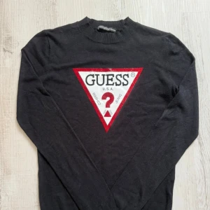 Svart långärmad tröja från Guess - Svart långärmad tröja från Guess med stort ikoniskt trekantslogo framtill i vitt och rött. Tröjan har rund halsringning och är tillverkad i mjukt bomullsmaterial. Perfekt för dig som gillar streetstyle och vill sticka ut med ett tydligt varumärke.