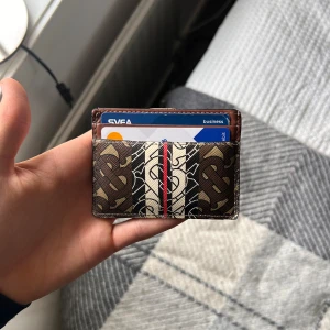 Burberry korthållare i skinn - Slimmad korthållare från Burberry i brunt och beige skinn med klassiskt monogrammönster och en röd rand i mitten. Perfekt för dig som vill ha stilren och lyxig känsla i fickan. Flera fack för kort och smidig att bära med sig.