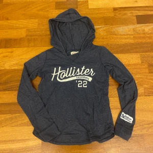 Mörkblå tunn hoodie från hollister - Mörkblå hoodie från Hollister med vit logga och text framtill. Tröjan har långa ärmar, huva och en liten vit detalj på ena ärmen. Perfekt för en avslappnad stil och enkel att matcha med jeans. Hoodien är lika tunn som en t skjorta!!
