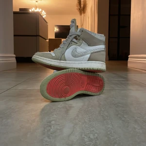 Nike Air Jordan 1 Zoom Air grå/vit - Nike Air Jordan 1 Zoom Air sneakers i high-modell med vit läderbas och ljusgrå mockadetaljer. Skorna har grå snörning, röd Zoom Air-logga på plösen och en sula med röd undersida. Perfekt för dig som gillar klassisk streetstyle med modern twist.