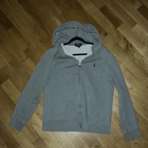 Grå hoodie från Polo Ralph Lauren - Snygg grå hoodie från Polo Ralph Lauren med dragkedja och huva. Tröjan har lång ärm och klassisk blå broderad logga på bröstet. Mjuk insida och ribbade muddar vid ärmslut och nederkant. Perfekt för chill dagar och enkel att matcha med jeans eller joggers. Lite små hål här och där, inget man märker