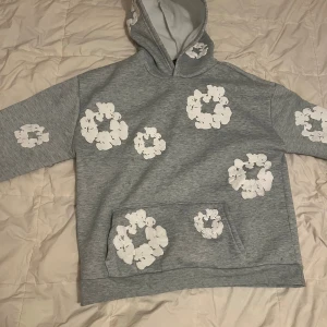 Grå hoodie med vita blommor Denim Tears - Grå hoodie från Denim Tears med stora vita blommor tryckta över hela tröjan, inklusive huvan och ärmarna. Tröjan har en klassisk känguruficka framtill och är tillverkad i mjukt bomullsmaterial. Perfekt för dig som gillar streetwear med unik design.