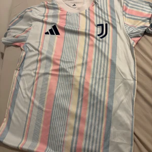 Juventus randig fotbollströja Adidas S träningströja - Juventus fotbollströja från Adidas i slim fit, storlek S. Tröjan har ett fräscht randigt mönster i ljusblått, rosa, vitt och ljusgult. Kortärmad modell med rund hals och Juventus-logga på bröstet. Tillverkad i lätt och ventilerande polyester.