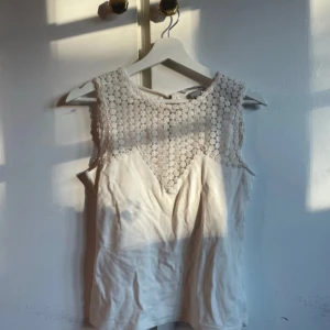 Beige ärmlös blus med spetsdetalj - Säljer en beige ärmlös blus med snygg spets upptill i geometriskt mönster. Blusen har rund halsringning och knäppning bak i nacken. Perfekt för dig som gillar romantiska detaljer och en ljus, fräsch look.