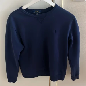 Marinblå sweatshirt från Polo Ralph Lauren - Snygg marinblå sweatshirt från Polo Ralph Lauren med diskret broderad logga på bröstet. Tröjan har rund hals, ribbade muddar och långärmad passform. Perfekt för dig som gillar klassisk stil med en modern touch.