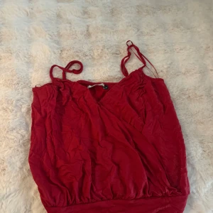 Röd  linnetopp med smala band - Säljer en snygg röd linnetopp med omlott-design och smala axelband som kan knytas. Toppen har en mjuk, lite glansig yta och ett brett band nedtill som ger en cool siluett. Perfekt för dig som gillar färg och vill sticka ut lite extra.