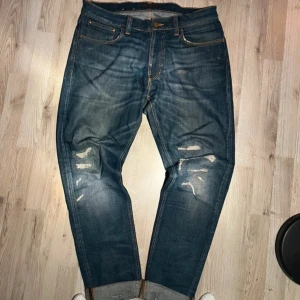 Blå jeans med slitningar från Nudie - Säljer ett par blå jeans från Nudie med coola slitningar och lagom tvättad look. Jeansen har klassisk femficksdesign, uppvikta benslut och kontrastsömmar på bakfickorna. Perfekt för dig som gillar en avslappnad och trendig stil.