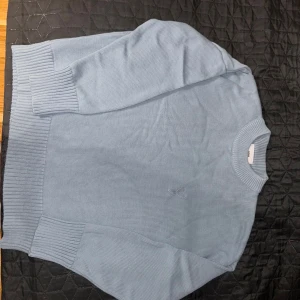 Ljusblå stickad tröja från Ami - Säljer en ljusblå stickad tröja från Ami Alexandre Mattiussi med broderad logga på bröstet. Tröjan har rund hals, breda ribbade muddar och är långärmad. Perfekt för dig som gillar stilren och clean design.