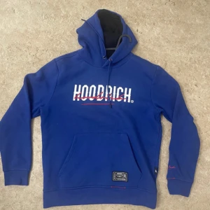 Blå HOODRICH hoodie med tryck - Snygg blå hoodie från HOODRICH med vit och röd logga på bröstet och ryggen. Tröjan har huva med snörning, känguruficka och coola detaljer på ärmen och nederkanten. Perfekt för dig som gillar streetwear och vill sticka ut.
