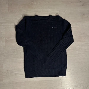 Mörkblå Bondelid - Snygg mörkblå kabelstickad tröja från Bondelid med diskret broderad logga på bröstet. Tröjan har rund hals, ribbade muddar och långärmad passform. Perfekt för dig som gillar klassisk stil med en modern touch.