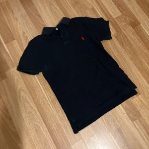 Svart pikétröja Polo Ralph Lauren - Svart pikétröja från Polo Ralph Lauren i slim fit-modell. Klassisk krage, korta ärmar och knäppning framtill. Röd broderad logga på bröstet ger en snygg kontrast. Tillverkad i mjuk bomull som är skön mot huden. Piken är lite mindre än S den sitter som 152 som motsvarar M i barnstorlek! Skriv till mig om pris eller om bilder!