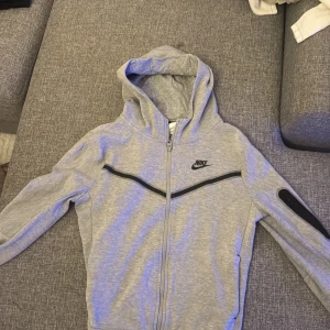 Grå Nike kofta med dragkedja - Snygg grå kofta från Nike med svart logga på bröstet och svarta detaljer längs dragkedjan och ärmen. Tröjan har huva, hel dragkedja och långa ärmar. Perfekt för chill eller träning, riktigt skön och stilren design.