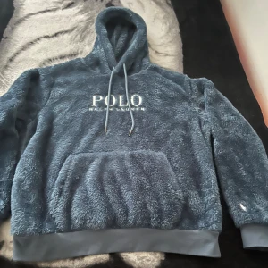 Blå fluffig hoodie från Polo Ralph Lauren - Supermjuk och fluffig blå hoodie från Polo Ralph Lauren med broderad logga på bröstet och ärmen. Tröjan har huva med snörning, stor magficka och ribbade muddar. Perfekt för kalla dagar när du vill vara både varm och snygg.