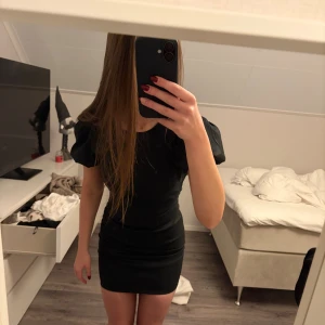 Svart kort klänning med puffärm - Snygg svart kort klänning med puffiga korta ärmar och figursydd passform. Klänningen har en  öppen rygg med knapp i nacken och dragkedja bak. Klänningen är aldrig använd.