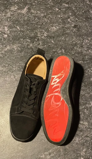 Loubs - Säljer ett par svarta sneakers från Christian Louboutin i mjuk mocka med klassisk rund tå och platt sula. Insidan är beige och yttersulan är röd med Louboutins signatur. Skorna har snörning och stilrena detaljer som passar dig som gillar lyxig streetstil.
