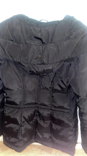 Roco Baroco - svart vinterjacka   - Svart vinterjacka från Roco Baroco med flera fickor, dragkedja och tryckknappar. Jackan har en hög krage och quiltade detaljer som ger en cool look. Materialet är glansigt syntet och jackan är långärmad, perfekt för kalla dagar under hösten och vintern!