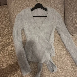 Ljusgrå fluffig omlottopp med knyt - Supermjuk och fluffig ljusgrå omlottopp med långa ärmar och breda knytband i midjan. Toppen har v-ringad hals och är stickad i ett lurvigt material som ger en cozy vibe. Perfekt för dig som gillar att sticka ut med en mysig look.