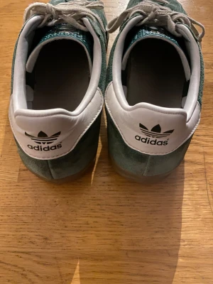 Adidas Gazelle gröna sneakers mocka - Säljer ett par Adidas Gazelle sneakers i grön mocka med vita detaljer och klassiska tre ränder på sidan. Storlek 44!!