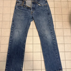 Levi's 501 blå jeans straight fit - Klassiska Levi's 501 jeans i blå denim med raka ben och normal passform. Jeansen har fem fickor, knappgylf och den ikoniska läderpatchen bak. Snyggt slitna detaljer och kontrastsömmar ger en tidlös look. Perfekta för dig som gillar autentisk vintagekänsla.