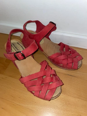Röda sandaler från Moheda - Röda sandaler från Moheda med flätade remmar i mocka och justerbart spänne vid ankeln. Skorna har öppen tå och en platt sula i trä-look.