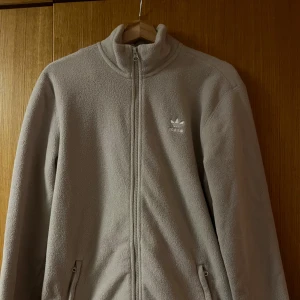 Beige fleecejacka - Säljer en beige fleecejacka från Adidas med dragkedja hela vägen och hög krage. Jackan har två fickor med dragkedja och Adidas-logga broderad på bröstet. Perfekt för kyliga dagar och har en clean, enkel look.