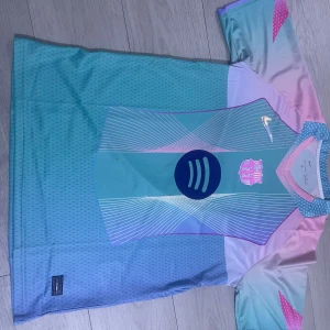 Barça x Nike pastell fotbollströja - Unik FC Barcelona fotbollströja från Nike i pastellfärger med blå, rosa och lila toner. Tröjan har korta ärmar, modern grafik och klubbmärke samt Nike-logga på bröstet. Tillverkad i lätt och ventilerande funktionsmaterial, perfekt för match eller träning.