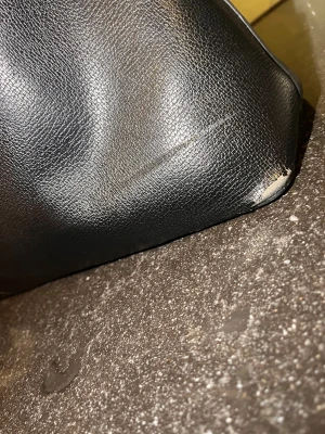 Svart handväska från Valentino - Stilren svart handväska från Valentino med guldfärgade detaljer och dragkedja. Väskan är tillverkad i skinnimitation och har rymligt foder med logotypmönster. Perfekt storlek för vardagens essentials och har dubbla handtag för enkel bäring. Lite slitage på nedre sidor, se bild.