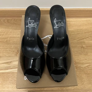 Svarta Louboutin pumps med röd sula - Säljer ett par ikoniska svarta pumps från Christian Louboutin med glansig finish och klassisk röd sula. Skorna har öppen tå, hög klack och är tillverkade i lackat skinn. Perfekt för dig som vill sticka ut med en lyxig och edgy look. De är replicas