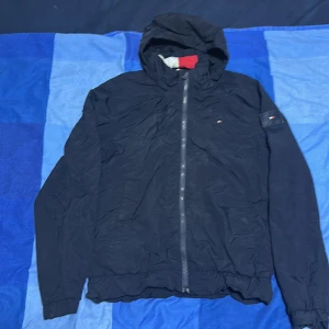 Svart vindjacka från Tommy Hilfiger - Svart vindjacka från Tommy Hilfiger med huva och hel dragkedja framtill. Jackan har diskret logga på bröstet och ärmarna, samt röd och vit detalj på insidan av huvan. Tillverkad i lätt polyester, perfekt för blåsiga dagar. Snygg och enkel design med elastisk nederkant.