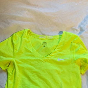 Neongul Nike Pro t-shirt med v-ringning - Nike Pro t-shirt i en riktigt snygg neongul färg. T-shirten har korta ärmar, v-ringning och en vit Nike-logga på bröstet. I träningsmaterial så perfekt vid hård träning
