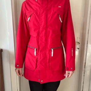 Röd Skaljacka Didriksons  - Klassisk röd skaljacka av märket Didriksons. Den har dragsko i midjan, nedtill och runt luvan så du kan välja hur du vill att den ska sitta. Det finns en innerficka och fyra framfickor. Tryck knappar ned till på armarna som kan fästas i två lägen. Stängs med dragkedja och tryck knappar. I fint skick och säljs då den inte används längre. Finns en knappt märkbar fläck framtill längre ner på jackan (se sista bild).