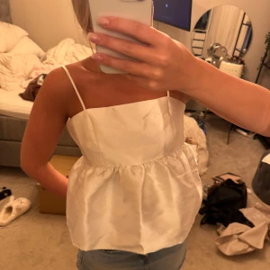 Vit siden linnetopp med smala axelband - Säljer en superfin vit linnetopp i glansigt sidenmaterial med smala axelband och lätt puffig, rynkad nederdel. Toppen har en rak halsringning och är perfekt för dig som gillar en clean och chic look. Passar snyggt till jeans eller kjol.