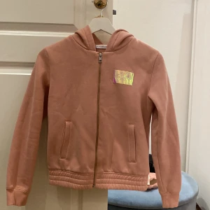 Rosa hoodie från Calvin Klein Jeans - Snygg rosa hoodie från Calvin Klein Jeans med huva och dragkedja framtill. Tröjan har ett holografiskt CK-märke på bröstet, två fickor och ribbade muddar vid ärmslut och nederkant. Perfekt för dig som gillar stilrena och bekväma plagg.