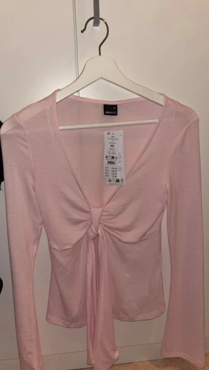Ljusrosa långärmad topp Gina Tricot - Supersöt ljusrosa långärmad topp från Gina Tricot med knyt framtill och v-ringning. Mjuk i materialet, Enkel men ändå trendig design som lyfter vilken outfit som helst.