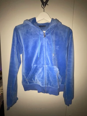 Blå velour hoodie Juicy Couture S - Säljer en blå hoodie från Juicy Couture i mjuk velour med broderad JC-logga på bröstet och en snygg J-dragkedja. Tröjan har huva med snörning, långa ärmar och känguruficka framtill. Perfekt för dig som gillar klassisk Y2K-stil! Ena metall ”pluppen” på snöret är borta men det är inget som märks.