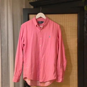 Rosa skjorta från Polo Ralph Lauren - Snygg rosa skjorta från Polo Ralph Lauren i custom fit. Skjortan har klassisk krage, knappar framtill och långärmad design. Tillverkad i mjuk bomull och har en liten blå logga broderad på bröstet. Perfekt för dig som gillar stilrena och färgglada plagg.