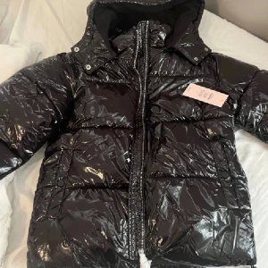 Svart glansig pufferjacka med huva - Säljer en svart glansig pufferjacka med huva och hög krage. Perfekt för dig som vill ha en varm och trendig jacka under kalla dagar. Knappt använd och hör av er om ni har några frågor