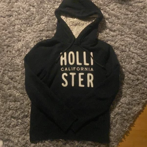 Blå  hoodie från Hollister med teddyfoder - Mysig blå hoodie från Hollister med stor vit broderad logga på bröstet. Insidan av huvan är fodrad med fluffigt teddyfoder i ljusgrått. Tröjan har snörning vid huvan och långa ärmar, perfekt för kalla dagar. Tröjan är i storlek L men passar mig som har M perfekt!! Vid minsta fråga är det bara att höra av sig!