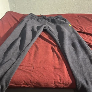 Svarta mjukisbyxor Lyle & Scott Junior - Svarta mjukisbyxor från Lyle & Scott Junior med elastisk midja och snörning. Byxorna har en gul logga på vänster sida och en bakficka. Mjukt material som är skönt att ha på sig, perfekt för chill eller träning.