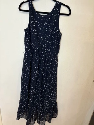 Marinblå långklänning med stjärnor H&M - Supersöt marinblå långklänning från H&M med vitt stjärnmönster. Klänningen är ärmlös, har rund halsringning och ett lätt rynkat midjeparti. Volangdetalj längst ner ger extra flow. Perfekt för dig som gillar en chill och lekfull stil.